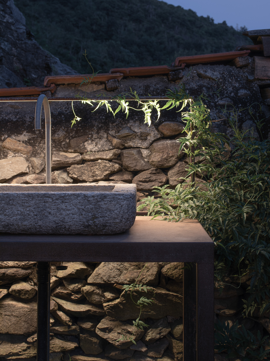 Dettaglio della cucina outdoor con vasca in pietra e luce integrata tra vegetazione e muratura storica.